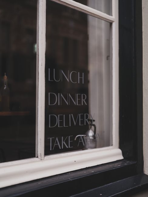 A close-up view of a white-painted window with several vertical panes, reflecting the dark interior of a flower shop. On the glass, white lettering advertises services including lunch, dinner, deliver
